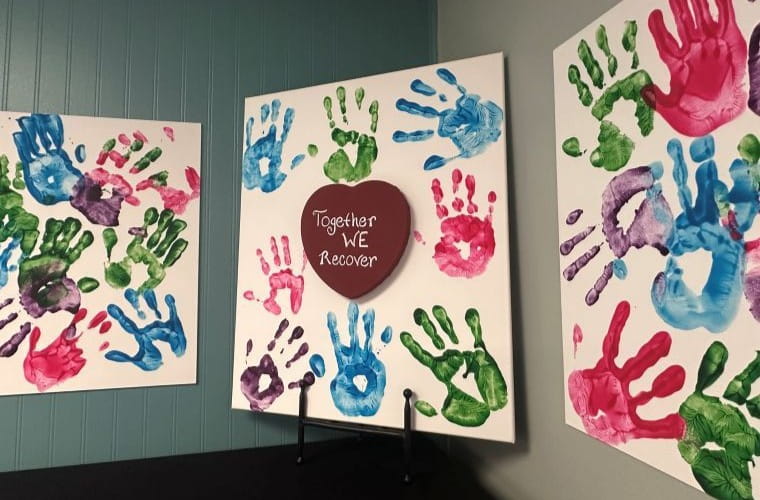 Colorful handprints in blue, green, red, and purple cover three white canvases on display. The center canvas features a heart-shaped cutout with the words "Together We Recover.