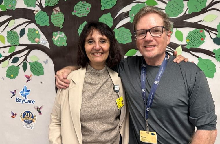 Two people stand smiling in front of a BayCare tree mural adorned with handwritten notes on green leaves. A BayCare logo and “20 years together” badge are visible. Both individuals wear name tags and one has an ID lanyard.
