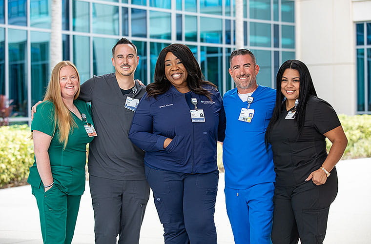 Three women and two men, each wearing a different color of medical scrubs, link arms and smile in front of a hospital building.
