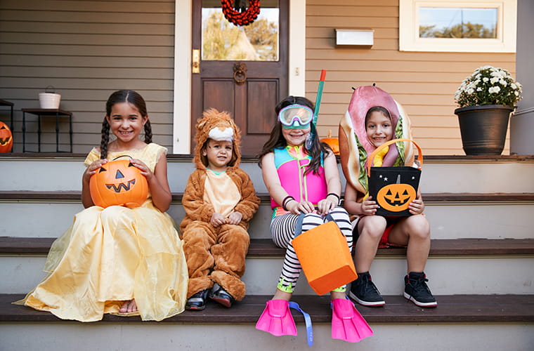 Four children in Halloween costumes sitting on porch steps: one in a yellow dress holding a pumpkin bucket, another in a lion costume, a third dressed as a diver with snorkel gear, and the last in a hot dog costume holding a pumpkin-shaped candy bag. Halloween decorations are visible. 