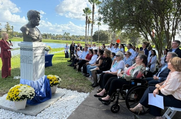 A speaker at a podium addresses seated attendees during an outdoor ceremony, with a bust of Hippocrates and floral arrangements displayed nearby.