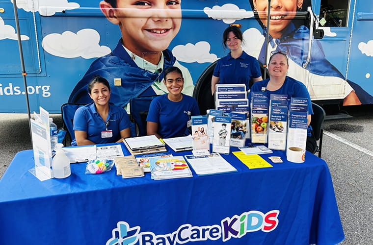 Four women are positioned behind a table covered with promotional materials under a blue canopy. The tablecloth displays the "BayCare Kids" logo. Behind them is a mobile unit decorated with children's images and cloud graphics. 