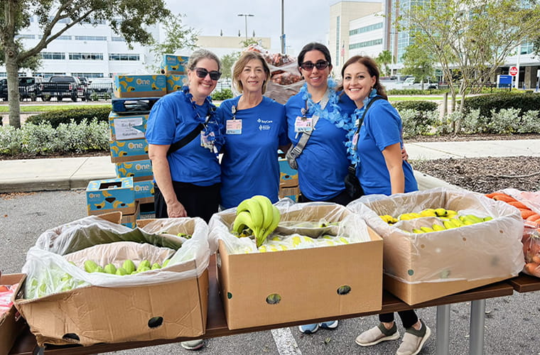 Four women wearing blue shirts stand behind a table filled with bananas and other produce. The event is taking place outdoors in a parking lot with trees and a large building visible in the background. 