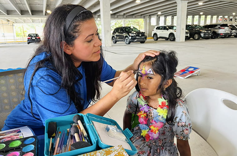 A person is painting a colorful butterfly design on a child's forehead at an outdoor event. Various painting supplies are visible on a table nearby. 