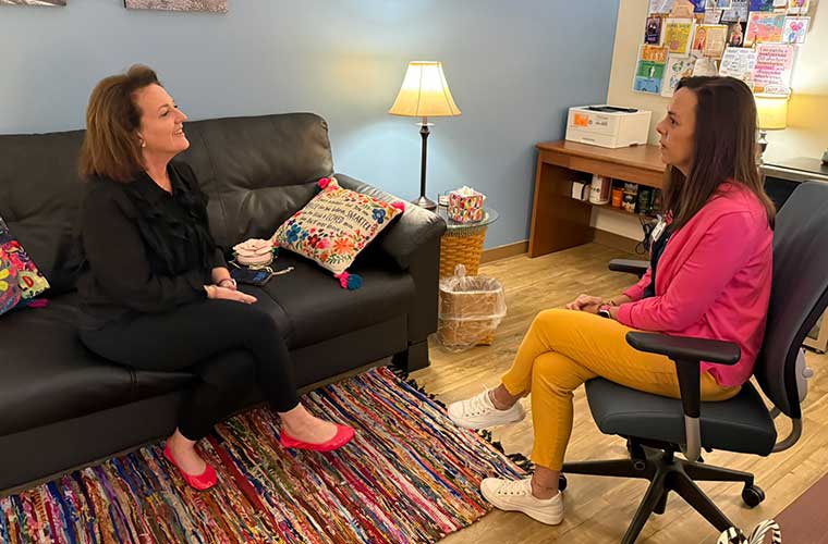 Two women sit across from each other in an office during a support therapy session for breast cancer patients. One woman listens while the other speaks.  