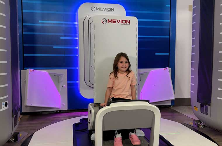A little girl wearing a T-shirt , pants and pink shoes sits in a medial device awaiting treatment.