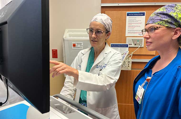 Two women dressed in hospital scrubs talk while looking at a computer screen.