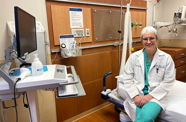 A woman dressed in surgical scrubs and a surgical cap and wearing glasses sits on a hospital bed with her legs crossed.