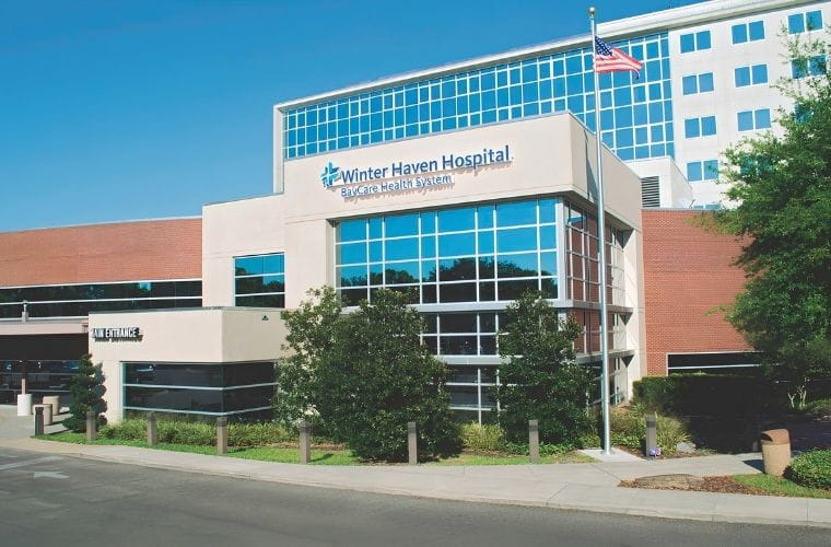 Exterior view of Winter Haven Hospital, a BayCare hospital, showing the main entrance, multi-story glass façade, hospital signage and an American flag flying in front of the building under a clear blue sky.