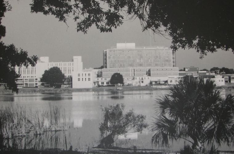 Black-and-white historic photograph showing Winter Haven Hospital from across a lake. The multi-story hospital complex stands on the opposite shoreline, with trees and vegetation framing the image in the foreground and around the edges. The hospital's reflection is visible on the calm water.