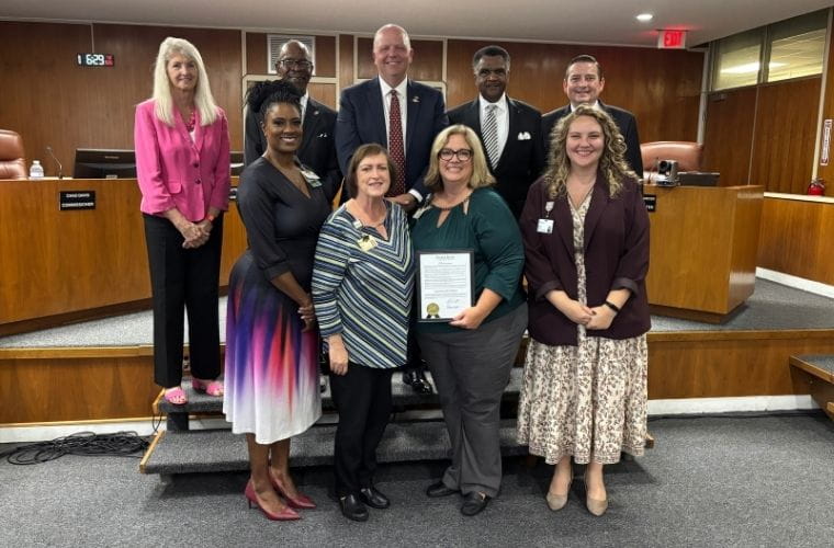 Four BayCare team members stand in the front row with members of the Winter Haven City Commission during a proclamation presentation in the City Commission chambers. One team member holds a framed proclamation signed by Mayor Brian Yates recognizing Winter Haven Hospital's 100 years of service to the community. The commissioners stand behind the BayCare representatives on the dais.