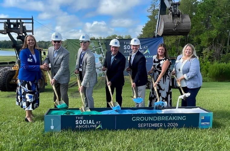 Seven people wearing hard hats participate in a ceremonial groundbreaking for The Social at Angeline on Sept. 1, 2026. Standing on a grassy site, the group holds gold-colored shovels filled with blue and green ceremonial dirt in front of a sign reading “The Social at Angeline Groundbreaking.” Construction equipment and trees are visible in the background.