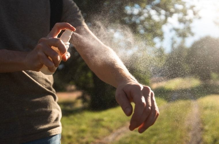 Person sprays insect repellent onto an exposed forearm while standing outdoors in a sunny setting.
