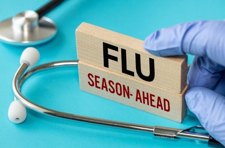 A stethoscope and a gloved hand holding wooden blocks that read "Flu Season Ahead" against a blue background.