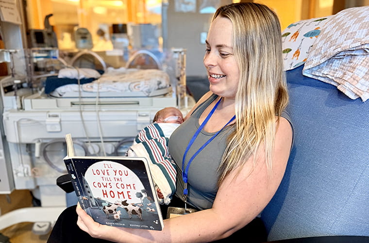 A woman holds a newborn in a hospital room while reading a baby book.