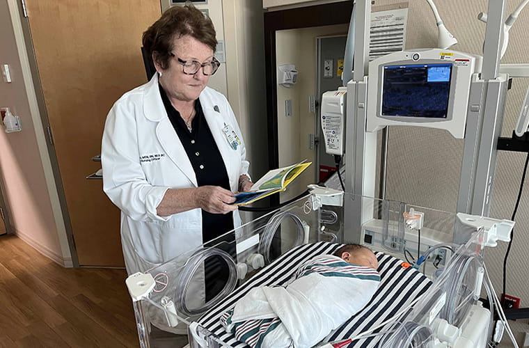 A woman in a white coat is reading to an infant lying in a clear hospital crib. The crib is in a medical room with various equipment nearby.