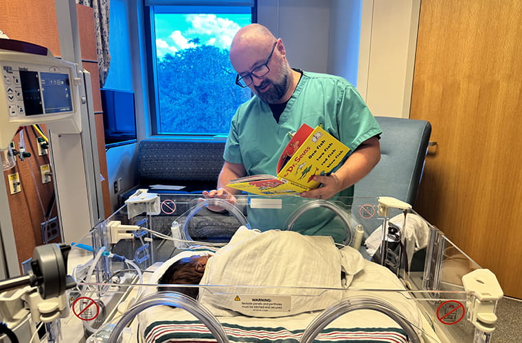A man in medical scrubs reads "Dr. Seuss's ABC" to a baby in an incubator in a hospital room.