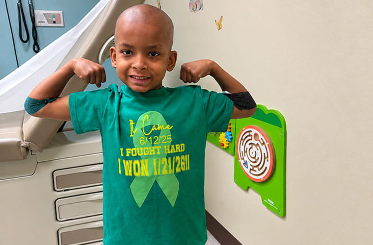 A smiling child, with a shaved head, stands in a hospital room flexing both arms. The child wears a green t-shirt with text: "It Came 6/12/25. I Fought Hard. I Won 1/21/26!!" The room features medical equipment and colorful wall decorations.