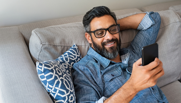 Happy smiling latin man using smartphone device while sitting on sofa at home