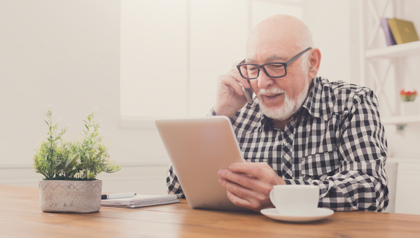 Senior man talking on phone and using tablet, sitting at desk at home