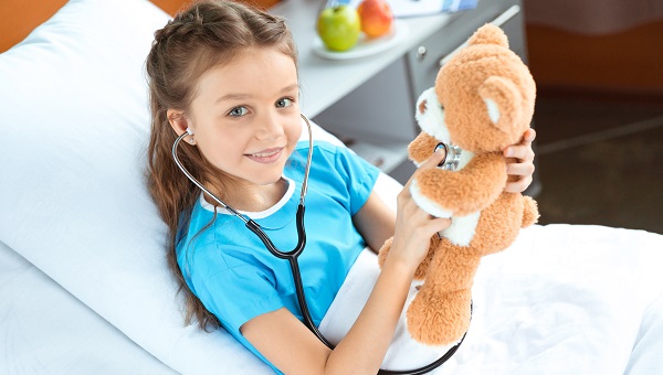 A girl in a hospital bed is using a stethoscope on her teddy bear.