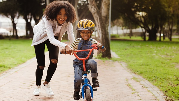 A mom is teaching her young son to ride a bicycle. He is wearing a helmet.