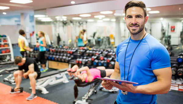 A personal trainer is holding a tablet while people are working out behind him.