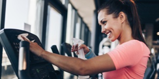 A woman is using disinfectant and a cloth to clean gym equipment.