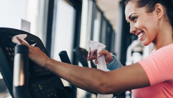 A woman is using disinfectant and a cloth to clean gym equipment.