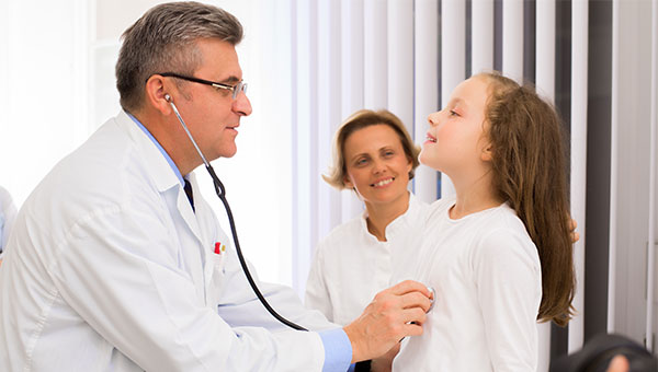 A mother watches a male doctor use a stethoscope to listen to the lungs of her young daughter