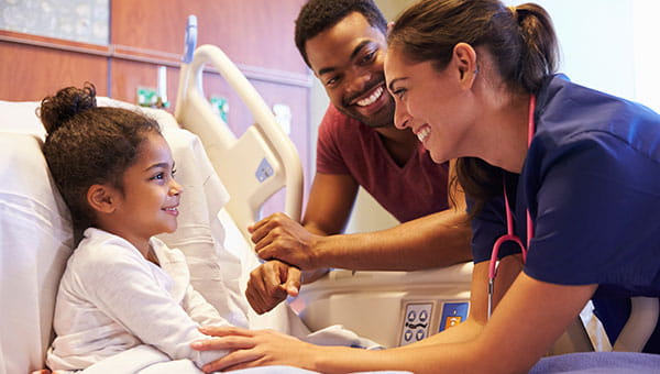 A smiling female nurse talks with a girl patient while her father stands next to her hospital bed