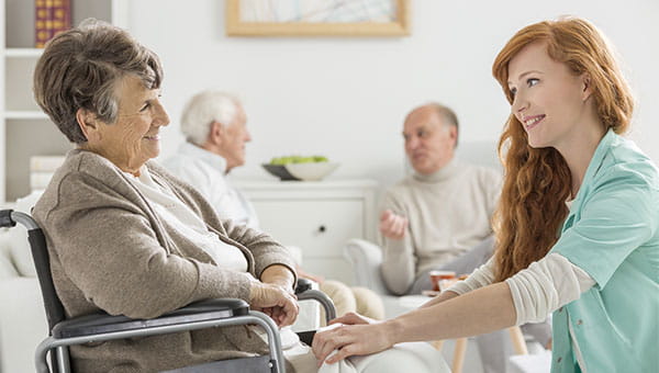 A smiling female nurse talks with a senior female patient who is sitting in a wheelchair.