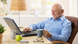 A senior man is sitting at a table, drinking coffee and looking at his laptop.