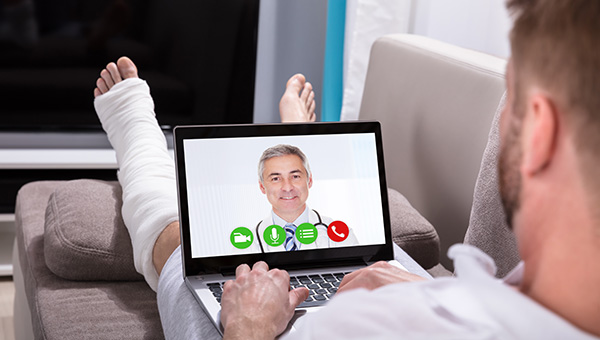 A man with a bandaged left foot, ankle and calf is resting on his sofa while talking to a doctor via video chat on his laptop.