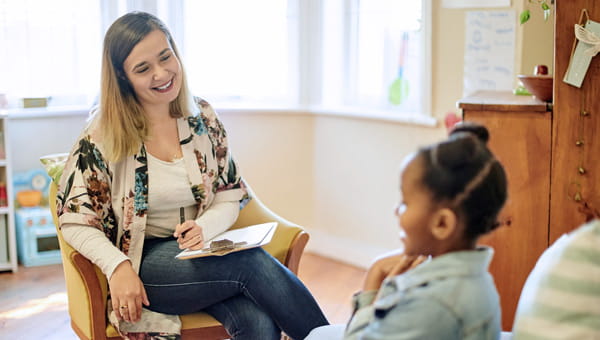 a smiling therapist with a clipboard and pen looking down at an African American child who seems to be talking