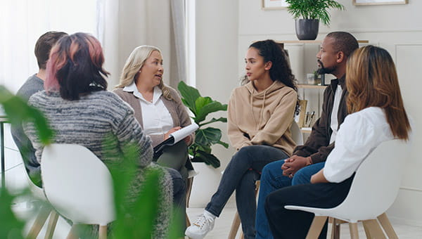 a group of 6 individuals sitting in a circle on white chairs, looking towards the facilitator 