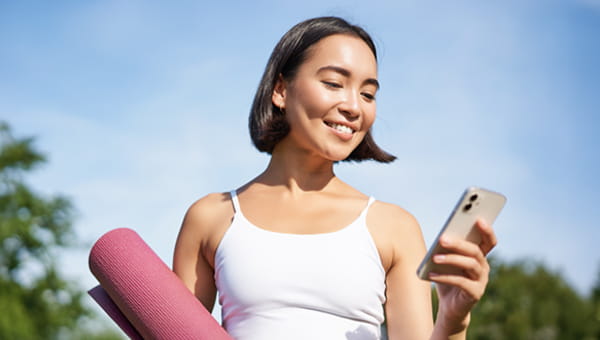 A smiling woman looking at her phone and carrying a yoga mat.