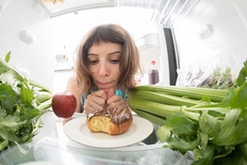 A woman is considering eating tasty healthy treats or a doughnut in her refrigerator.