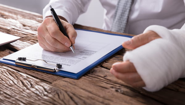 A man is completing a form and writing with his right hand while he rests his left arm, which is in a cast, on the table.