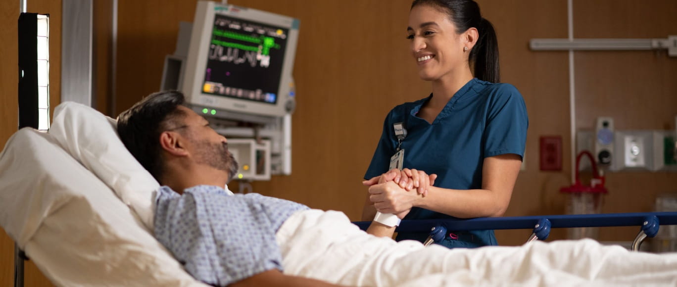 A smiling nurse holds the hand of a patient in a hospital bed.