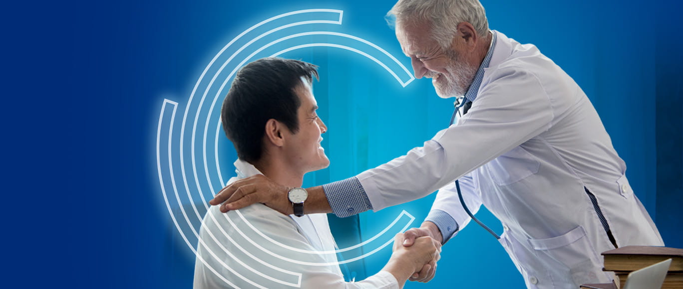 an Asian patient looking up while sitting shaking hands with an older white haired Caucasian male physician looking down at him