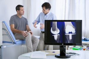 A male patient is sitting holding his knee while a  doctor speaks with knee X-rays in front of both them