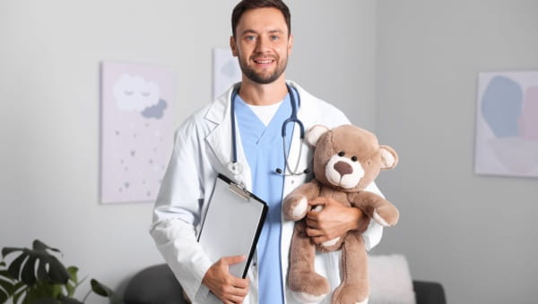 A Caucasian doctor soft smiling straight at the camera, holding a clipboard in one hand and a teddy bear in the other.