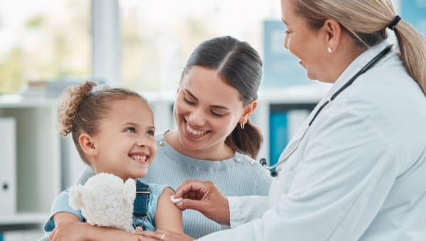 a female blonde doctor placing a cotton ball on a child's arm, the child is about 6 years old and smiling up at the doctor while holding a stuffed animal. The child is on her mom's lap, the mom is smiling looking at her child.