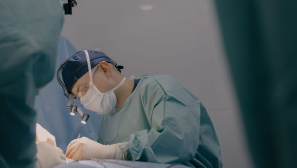 a male doctor in teal scrubs leaned over with a mask and a head lamp performing surgery
