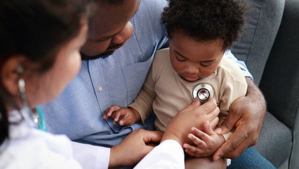 A physician listens to the heartbeat of a male toddler patient while his father holds him.