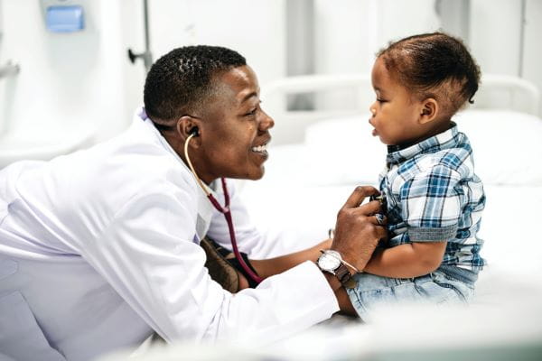 A smiling doctor listening to the heart of a toddler patient.