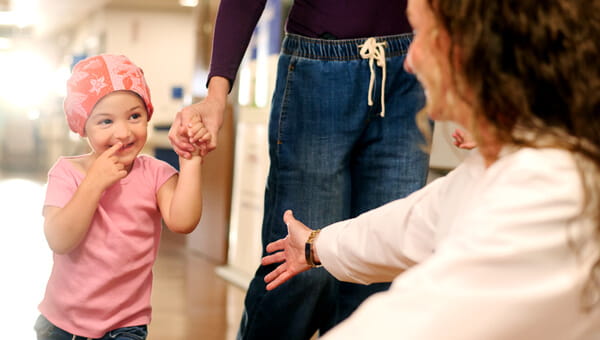 A little girl, who is a pediatric patient, is wearing a bandana and is walking up to a doctor while her mother is holding her hand