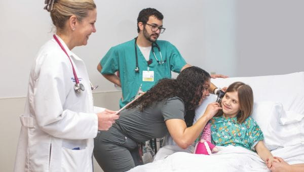A little girl, who is a pediatric patient is being examined by a group of doctors in her hospital bed, while one of the nurses is looking inside her ear with a medical tool