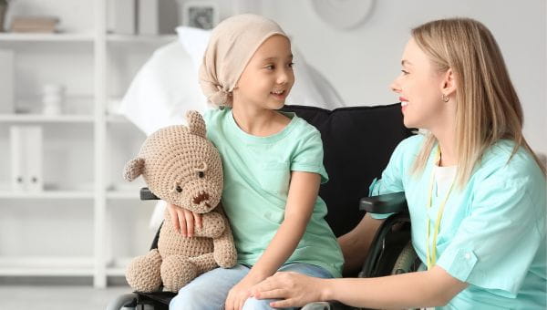 A female pediatric cancer patient holds a teddy bear while looking at a female nurse.